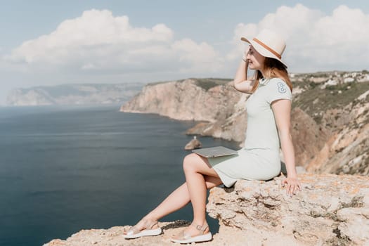 A woman in a straw hat sits on a cliff, holding a laptop, overlooking the vast blue ocean and rocky coastline.
