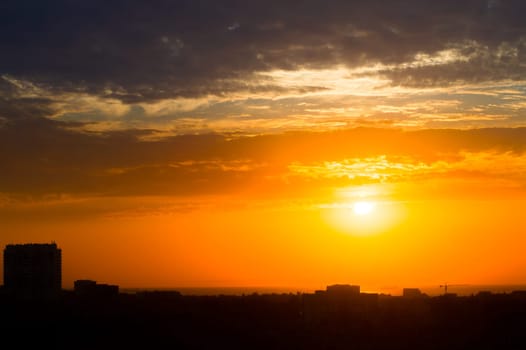 Dawn in a cloudy sky over a seaside town. The sun breaks through the clouds above the rooftops. Silhouettes of buildings and ships at anchor
