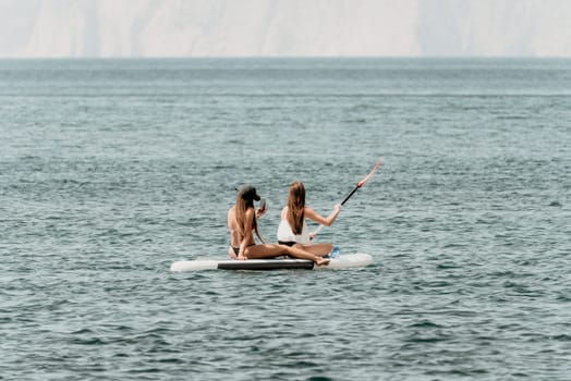 Two women are on a surfboard in the ocean. One of them is wearing a white shirt and the other is wearing a black bikini
