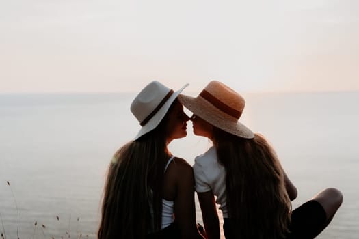 Two women wearing straw hats gaze at each other while sitting near the ocean at sunset.