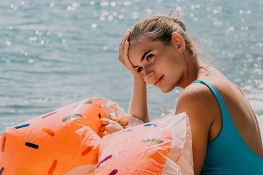 A woman wearing a blue swimsuit relaxes on a beach with colorful inflatable rings near a blue ocean.