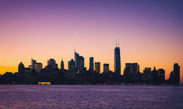 The Chicago skyline is visible at sunset. The tall buildings of the city are silhouetted against a sky that is a mix of orange, yellow, and purple. The moon is visible in the upper left of the image.
