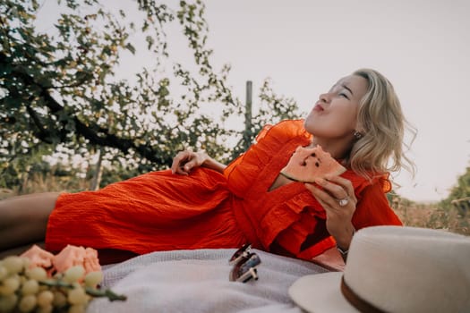 A woman in a red dress lays on a blanket in a grassy field, taking a bite of watermelon.