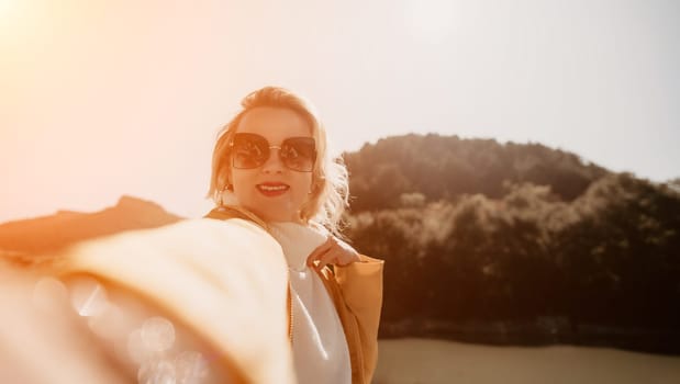 A woman in sunglasses takes a selfie while wearing a white dress with a mountain range in the background. The sun is shining brightly.