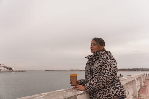 A woman in a leopard print coat is holding a coffee cup and looking out at the water