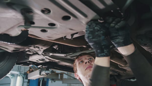 Car service workshop - mechanic unscrews detail of car under bottom of vehicle, close up