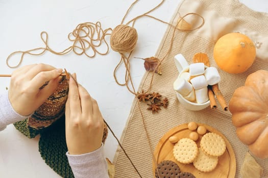 Hands knitting with yarn, surrounded by autumn snacks like cookies, marshmallows, and pumpkins for a cozy vibe.