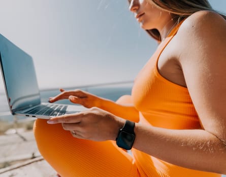 A woman wearing an orange outfit is sitting outdoors, using a laptop. She is likely working or browsing the internet.