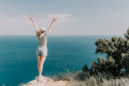 A woman with her arms raised in the air stands on a cliff looking out at the deep blue sea, capturing a sense of liberation and joy.