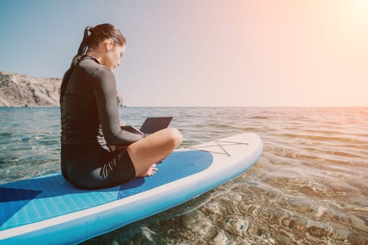 Digital nomad, woman in the hat, a business woman with a laptop sits on the rocks by the sea during sunset, makes a business transaction online from a distance. Freelance, remote work on vacation.