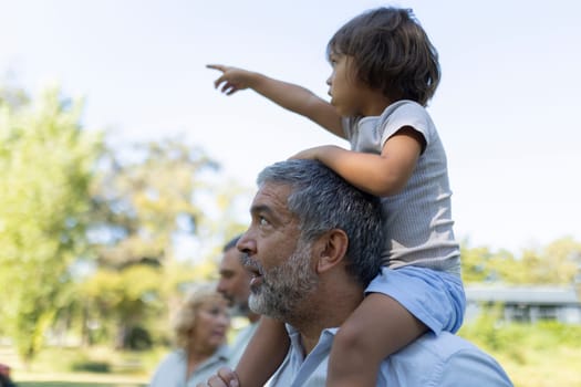 Little girl is enjoying a day at the park with her family, sitting on her grandfather's shoulders and pointing at something in the distance