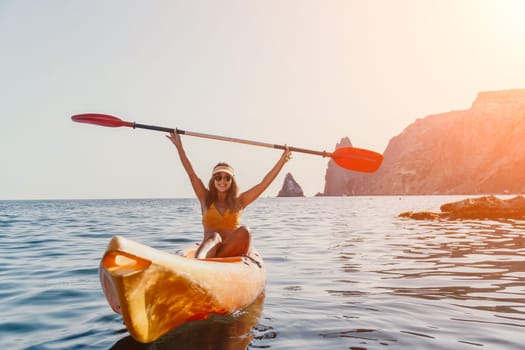 A woman in a bikini paddles a kayak with raised arms, enjoying a scenic ocean view with mountains in the distance.