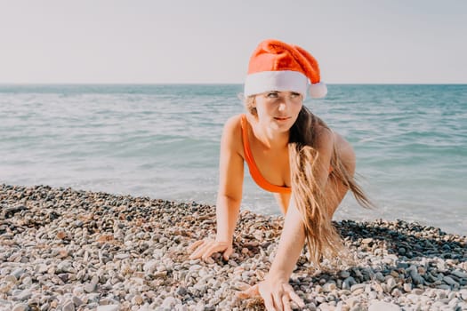 A woman wearing a red Santa hat and a bright orange bikini crawls along a pebble beach by the sea.