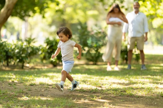 Little boy having fun running towards camera in a park while his parents are watching him in the background