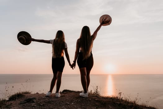 Two women in black shorts and white sneakers stand with their backs to the camera on a cliff, holding hands. They face the ocean at sunset and are silhouetted against the sky.