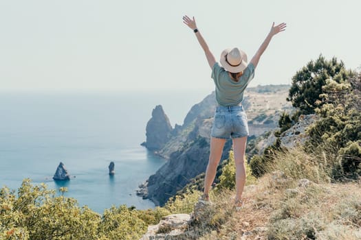 A woman in a hat and shorts stands with arms raised, looking out at the ocean from a clifftop, capturing the feeling of freedom and awe.