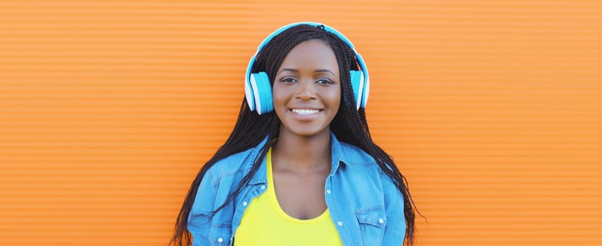 Portrait of happy African young woman listening to music with headphones, cool modern black teenage girl smiles on colorful bright orange background, summer day
