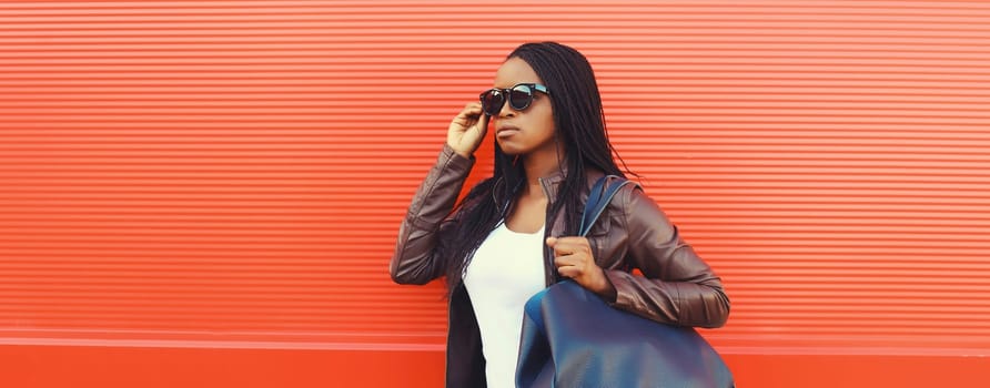 Fashionable young African woman with handbag in casual clothing, stylish modern black girl with dreadlocks, glasses posing in the city on red background