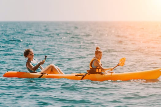 Two young women in a bright orange kayak paddle on a calm turquoise sea on a sunny day. The woman in the back is holding a paddle and the woman in the front is holding a selfie stick.