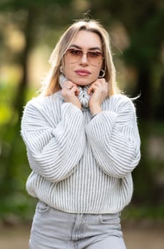 Blonde young woman is posing outside in a park wearing a turtleneck sweater and sunglasses