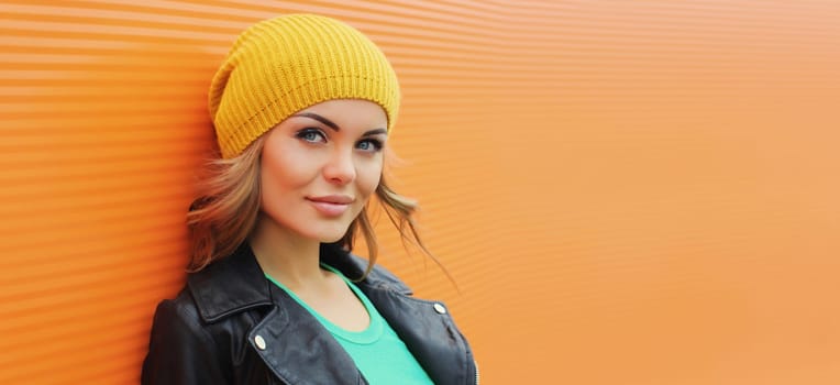 Portrait of beautiful young blonde woman posing in yellow hat, black leather jacket looking at camera on colorful orange background