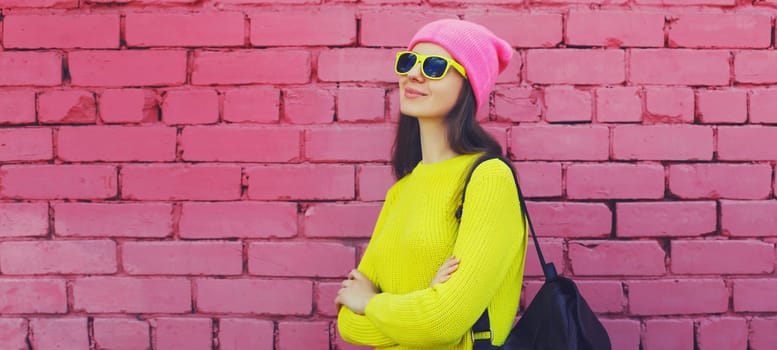 Stylish young teenager girl with backpack posing in vivid colorful clothes, yellow sweater, hat, glasses on city street on pink brick wall background