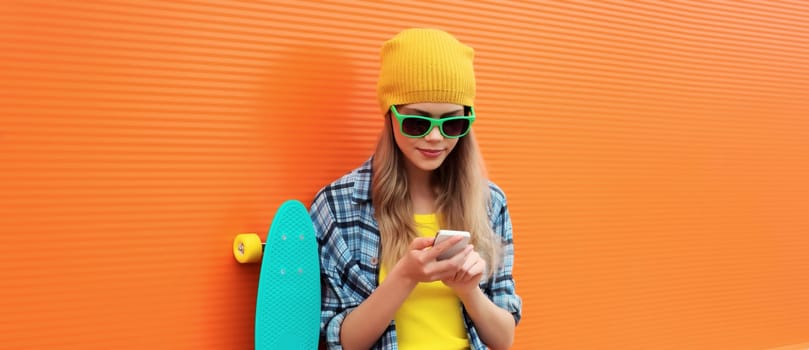 Modern stylish young woman using smartphone, teenage girl holding phone in colorful clothes with skateboard on city street on orange wall background