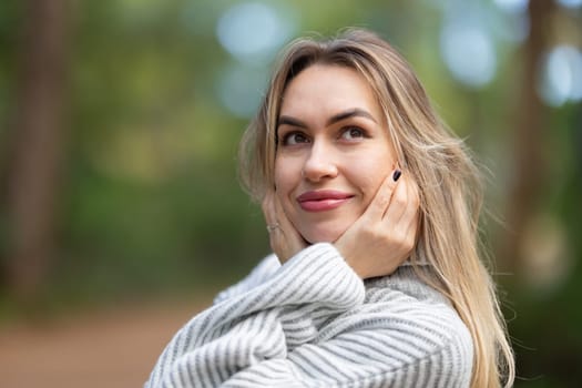 Young woman wearing a wool sweater is holding her head in her hands, daydreaming in a forest