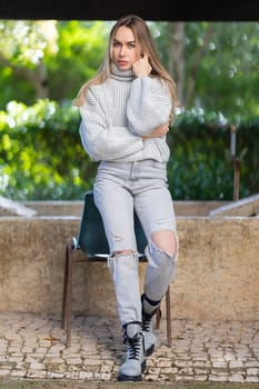 Young blond woman with long hair wearing jeans and a white sweater, posing sitting on a chair in a park