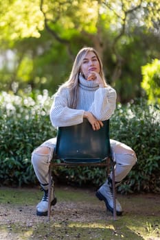 Young woman is posing sitting on a chair in a park, she is wearing a white sweater and ripped jeans