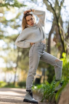 Young woman is posing in a park, touching her hair, wearing jeans and a sweater