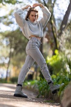 Young woman is standing on a path, holding her hair with her hands and wearing a wool sweater, jeans and boots