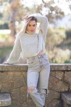 Attractive blond woman is posing touching her hair with one hand while leaning on a stone wall in a park
