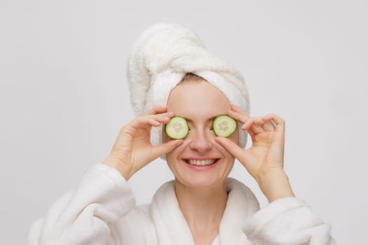 A woman in a white bathrobe and towel on her head holds cucumber slices over her eyes, smiling. The background is plain and light-colored, creating a spa-like atmosphere.