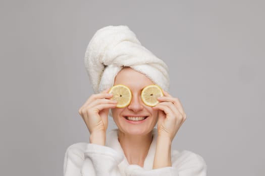 A happy girl in a towel and robe holds two lemon slices in her hand, covering her eyes with an orange. Skin care and moisturizing concept