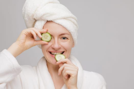 A woman in a white bathrobe and towel on her head holds cucumber slices over her eyes, smiling. The background is plain and light-colored, creating a spa-like atmosphere.