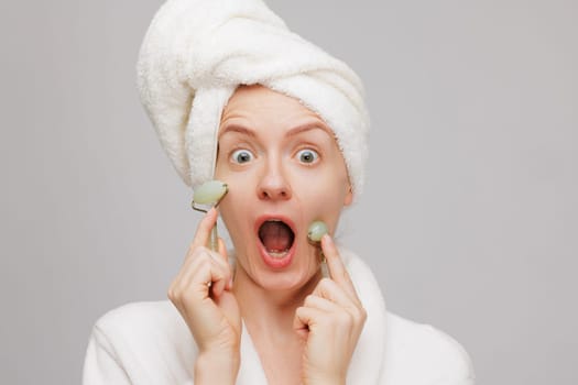 A surprised woman with a towel on her head and a bathrobe, holding jade rollers on her cheeks. She has wide eyes and an open mouth, expressing excitement or shock.