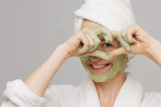 A woman with a towel on her head smiles as she applies a green face mask. She has a happy expression on her face, showing off her skincare routine