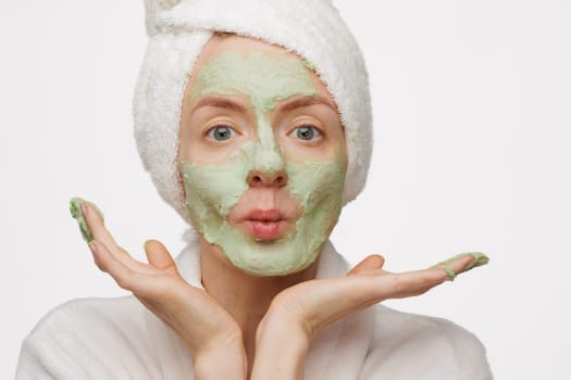 A woman with a towel on her head smiles as she applies a green face mask. She has a happy expression on her face, showing off her skincare routine