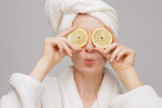 Emotional young woman in white towel on head and with facial mask and halves of ripe lemon on white background. Photo of woman receiving spa treatments. Beauty and Skin care concept