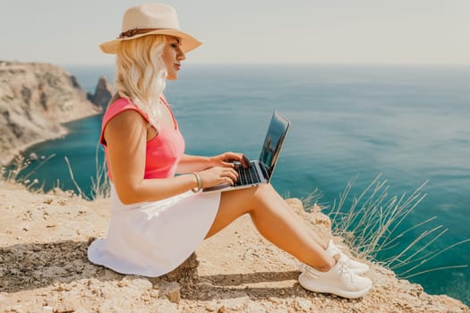 A woman is sitting on a rocky beach with a laptop open in front of her. She is wearing a pink top and a straw hat. Concept of relaxation and leisure