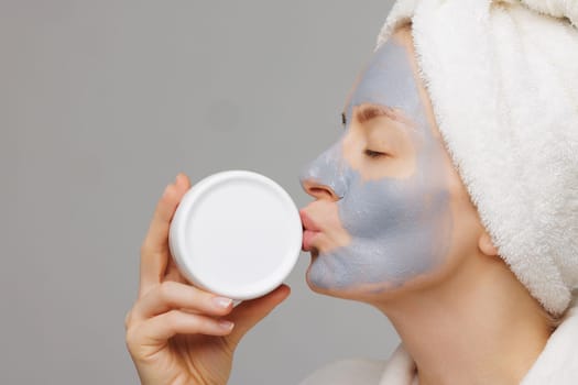 Woman with mask on her face playfully kisses her favorite white jar of cream. concept of skin care for face and body