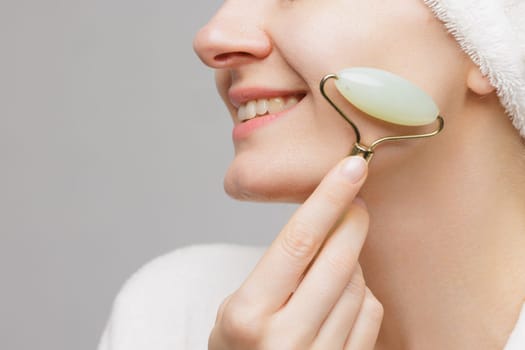 A woman in a white robe and a towel on her head smiles while holding jade facial rollers under her eyes.. wrinkle smoothing concept