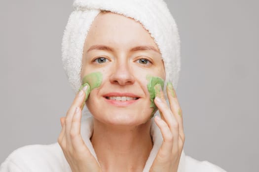 A woman with a towel on her head smiles as she applies a green face mask. She has a happy expression on her face, showing off her skincare routine