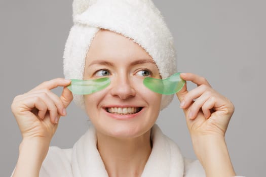 A woman with a towel on her head and a robe smiles as she applies green gel patches under her eyes facial cosmetology concept