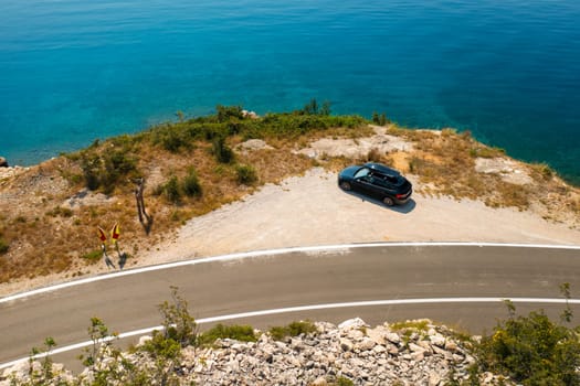 Car with a roof box, parked near a rugged cliff road, with the azure sea below surrounded by jagged rocks
