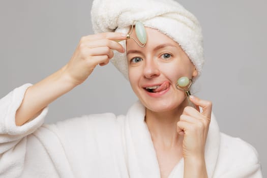woman with towel on head and robe holding jade rollers on cheeks sticking out tongue, wrinkle smoothing concept