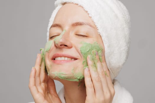 A woman with a towel on her head smiles as she applies a green face mask. She has a happy expression on her face, showing off her skincare routine
