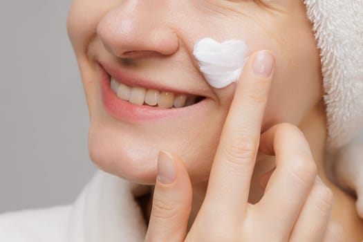 A woman applies cream to her cheek, smiling. She has a towel on her head and a robe on. The cream on her cheek is shaped like a heart.