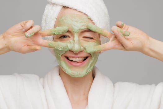 A woman with a towel on her head smiles as she applies a green face mask. She has a happy expression on her face, showing off her skincare routine